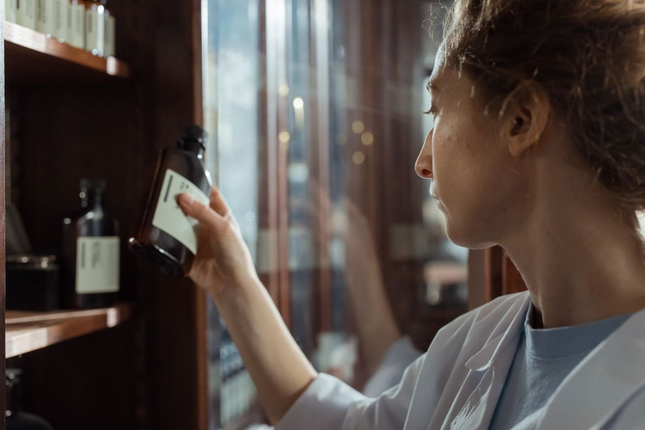 services-01 A pharmacist in a lab coat selecting a medication bottle from a pharmacy shelf indoors.